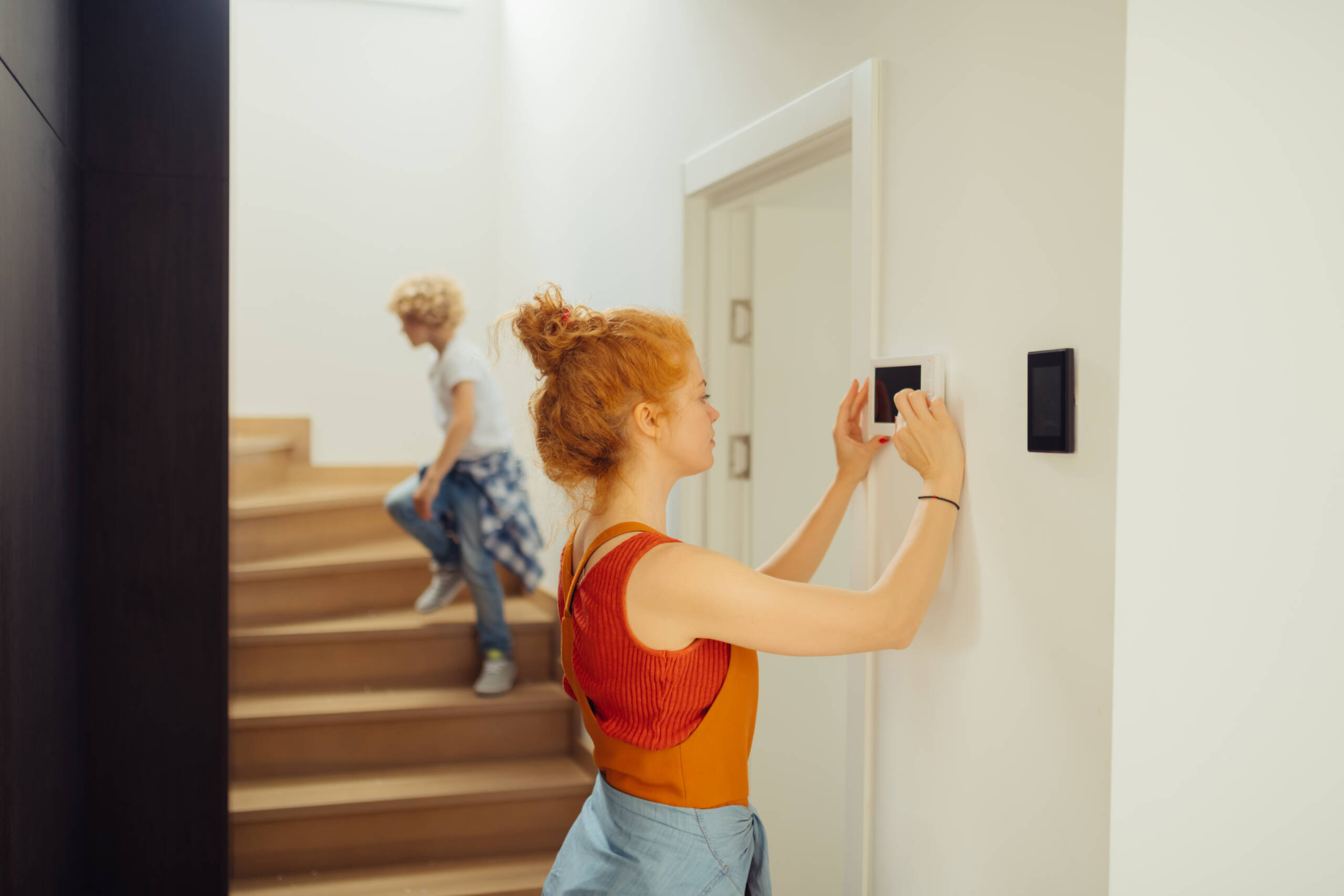Woman adjusts the thermostat of her home. A child can be seen running up the stairs in the background.