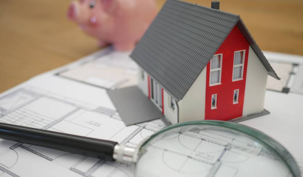 On a table sits an architectural map with a magnifying glass, a mini-house model and a piggy bank in the background.