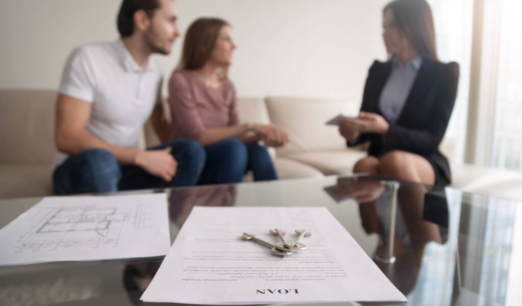 In the background there is a happy young couple discussing their mortgage with an estate agent. In the foreground of the image, a set of keys sits on a contract which the young couple have just signed.